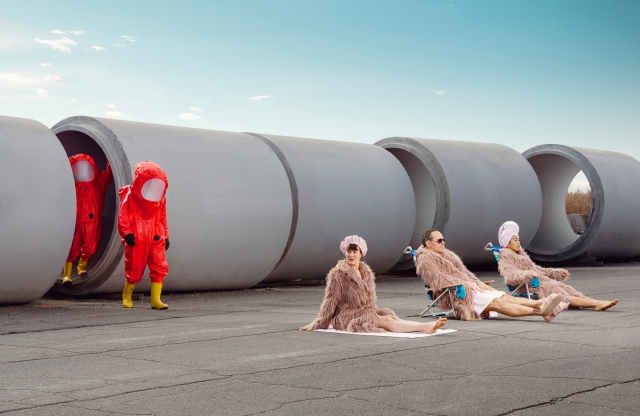From behind a row of giant concrete pipes, two hazmat-suit-clad figures approach a group of furry-robe-wearing sunbathers.