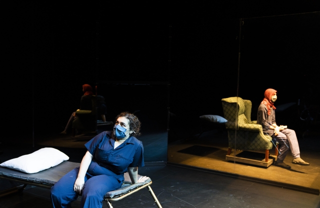 A masked woman sits on a cot looking off stage, while on the other side of a stage another masked woman with a headscarf looks in a different direction